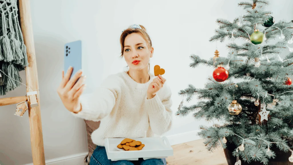 A woman in a white sweater takes a selfie while holding a heart-shaped cookie, sitting by a decorated Christmas tree with colorful ornaments.