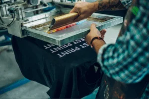 A man working on a printer, making a test print on a black t-shirt using screen printing.