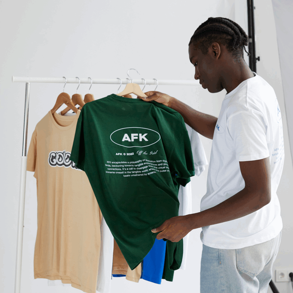 A man with braided hair examines a green t-shirt with white text on a clothing rack, alongside tan, white, and blue shirts.