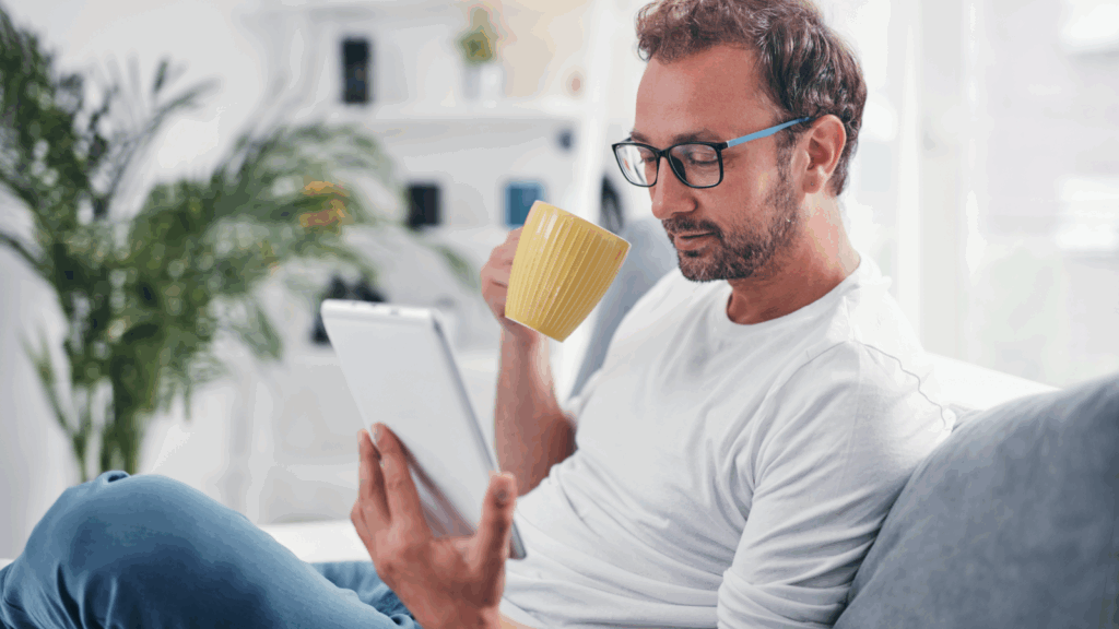A man with glasses, seated on a sofa, sips from a yellow mug while attentively reading a tablet.