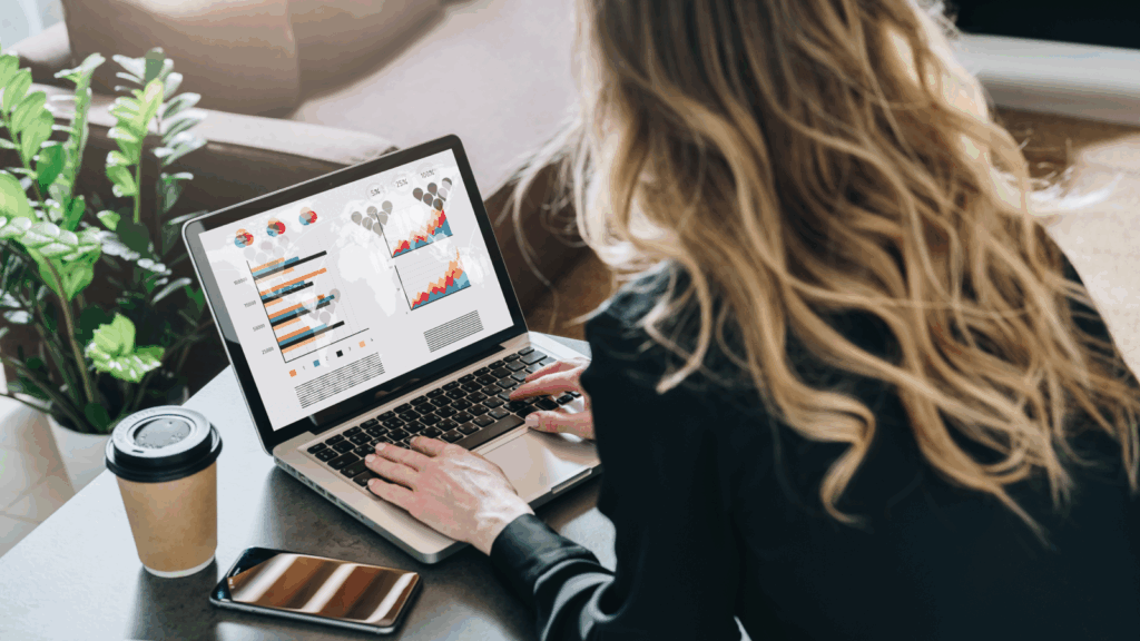 A woman works on a laptop displaying colorful charts and graphs, with a coffee cup, smartphone, and potted plants nearby.