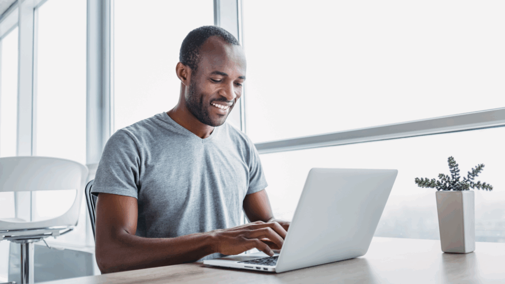 A smiling man sits at a table, working on his laptop with a relaxed demeanor.