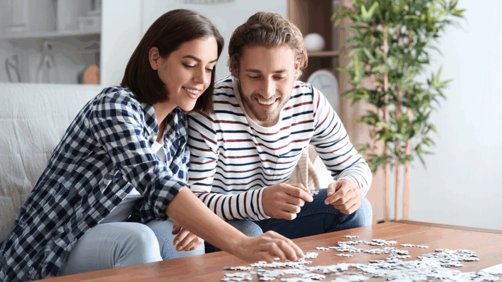 A smiling couple assembles a puzzle at a wooden table indoors. The woman wears a checkered shirt, and the man wears stripes.