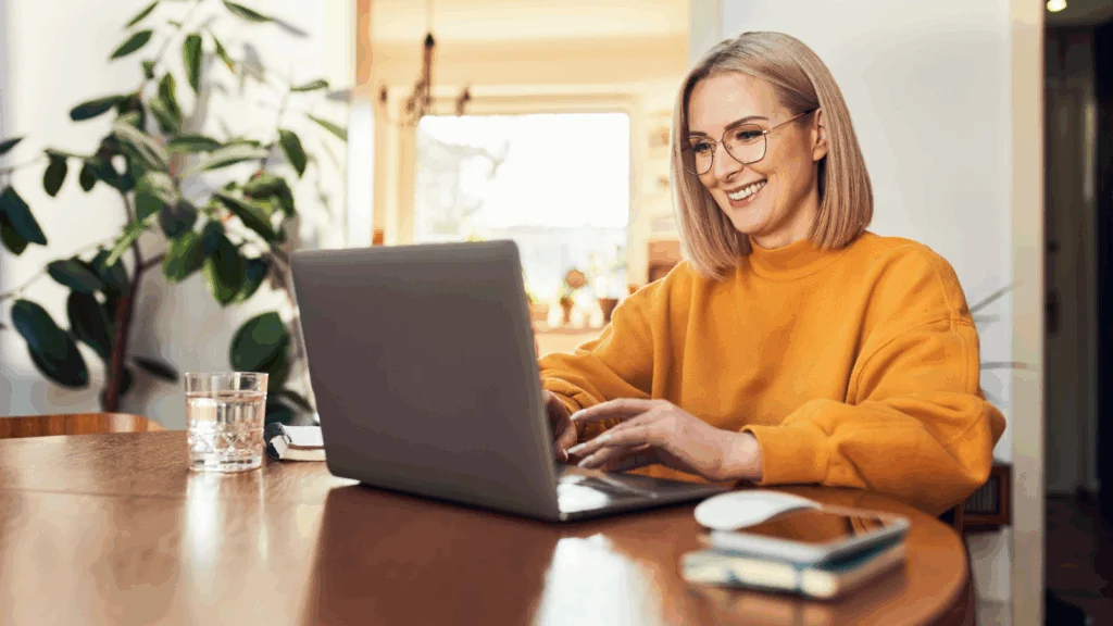 A woman wearing glasses sits at a table, focused on her laptop while working or studying.