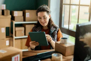 A woman wearing glasses smiles while using a tablet at her desk. Nearby are cardboard boxes and packing tape.