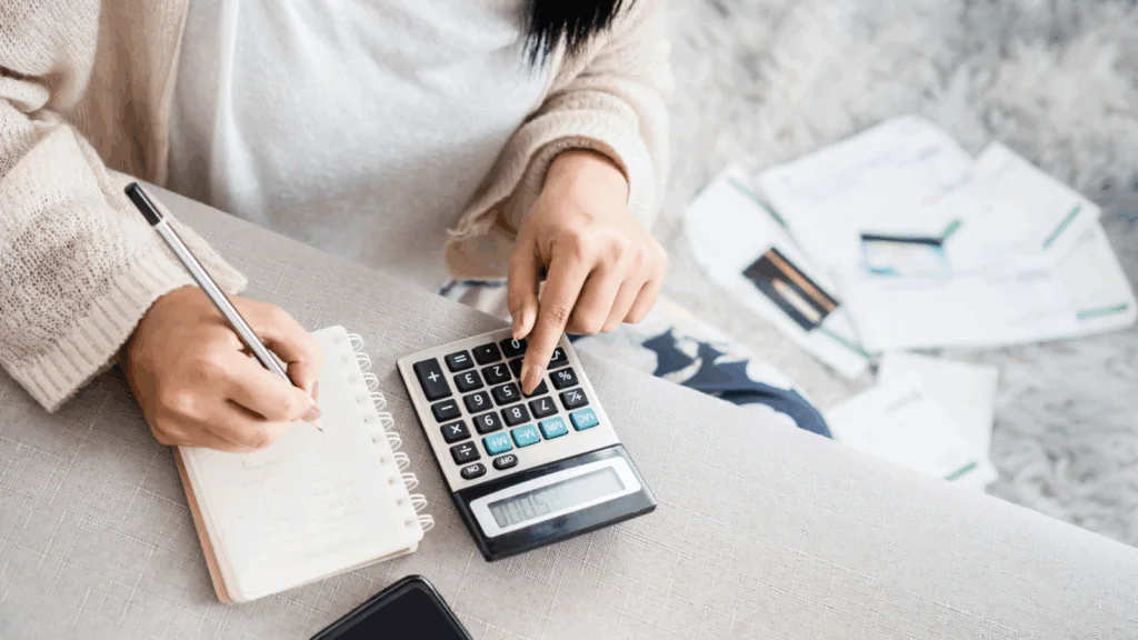 01 What are the main factors that affect profitability A woman uses a calculator and takes notes in a notebook. Bills and documents scatter on a soft rug.