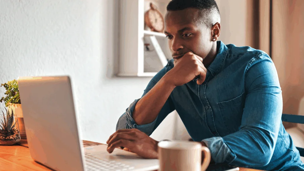 02 How to calculate your profit A man in a denim shirt sits at a wooden table, focused on a laptop, reading whether print-on-demand is profitable or not.
