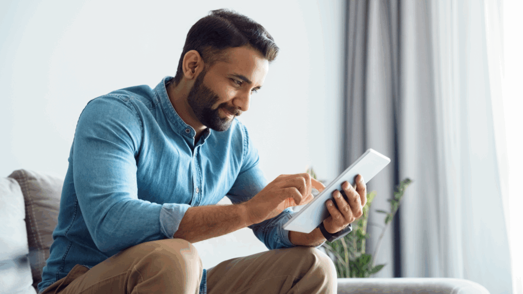 03 How to make your print on demand business more profitable A man in a blue shirt sits on a sofa, smiling and using a tablet to find out if print-on-demand is profitable.