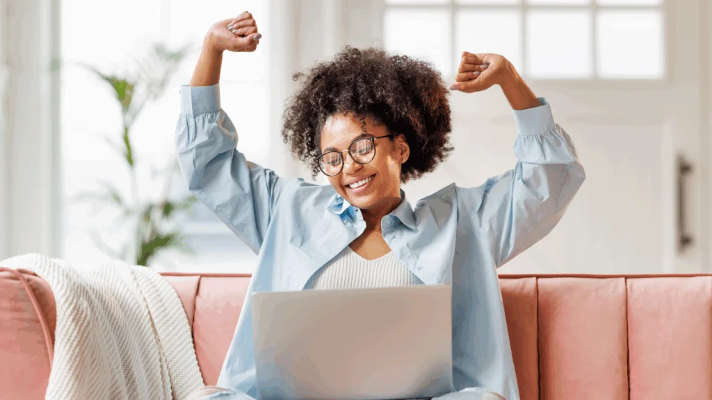 04 POD success stories A joyful woman with curly hair and glasses raises her arms in celebration while sitting on a pink sofa, using a laptop.