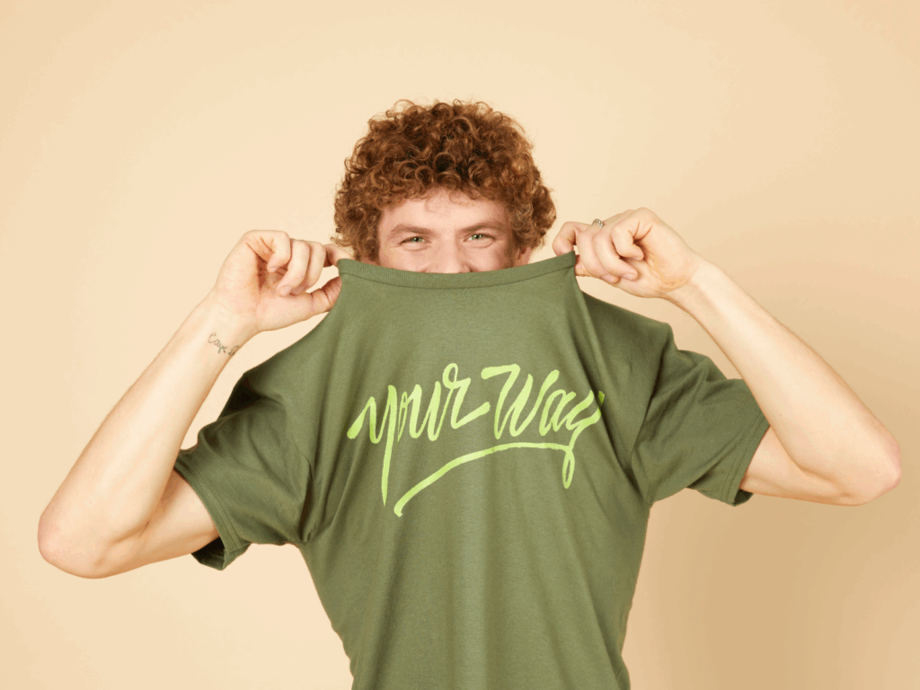 05 Start a print on demand business with Printify A smiling man playfully lifts their green t-shirt to expose their eyes. The shirt reads "your way" in light green script.