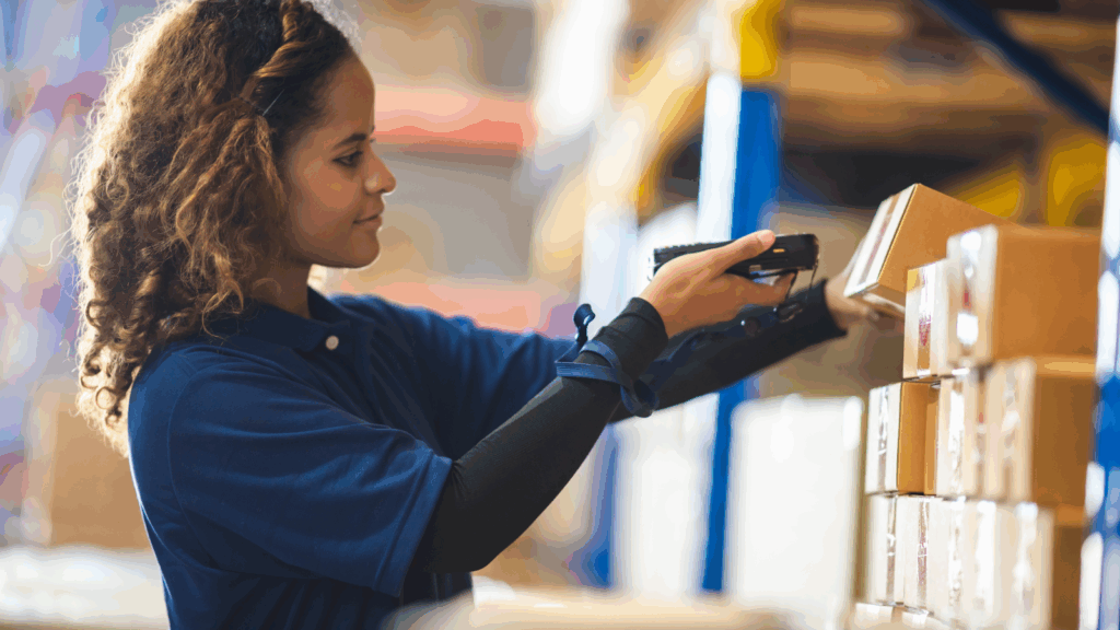 A woman in a warehouse holds a barcode scanner, focused on her task amidst shelves of inventory.