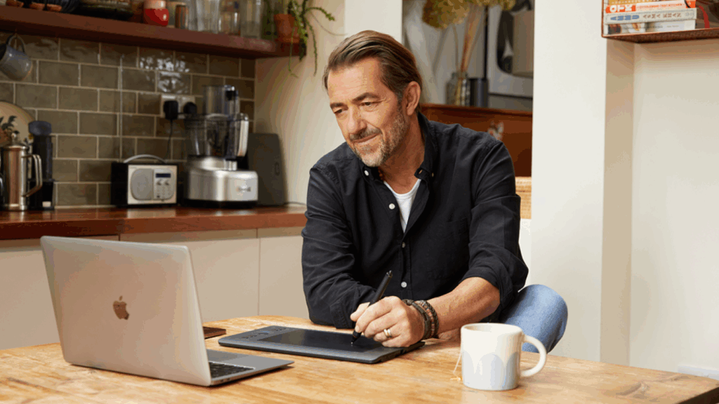 A man in a black shirt works at a kitchen table with a tablet and laptop, searching for how to sell supplements.