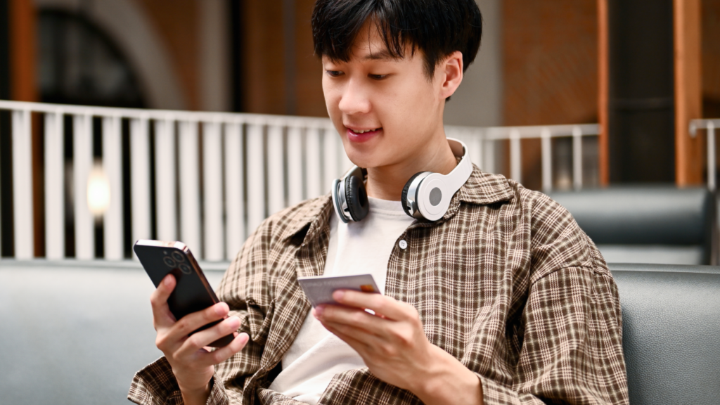 A young man with headphones around his neck sits indoors, using a smartphone and holding a credit card to pay for TikTok Promotion.