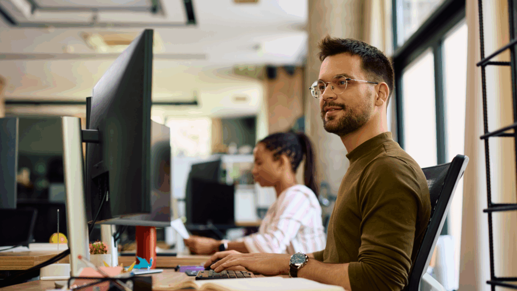 A man wearing glasses and a green shirt smiles while working at a computer in an office, learning how to sell coffee online.
