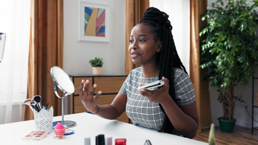 Brand and sponsor deals A woman with braided hair in a plaid dress sits at a table with makeup items and a mirror, recording herself using a phone.