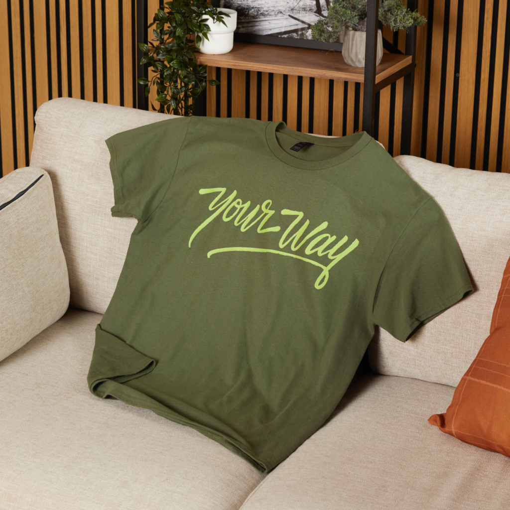 Bright color palettes and minimalist prints Green t-shirt with "Your Way" in yellow cursive font rests on a beige sofa. A small plant and decor items sit on a wooden shelf behind it.