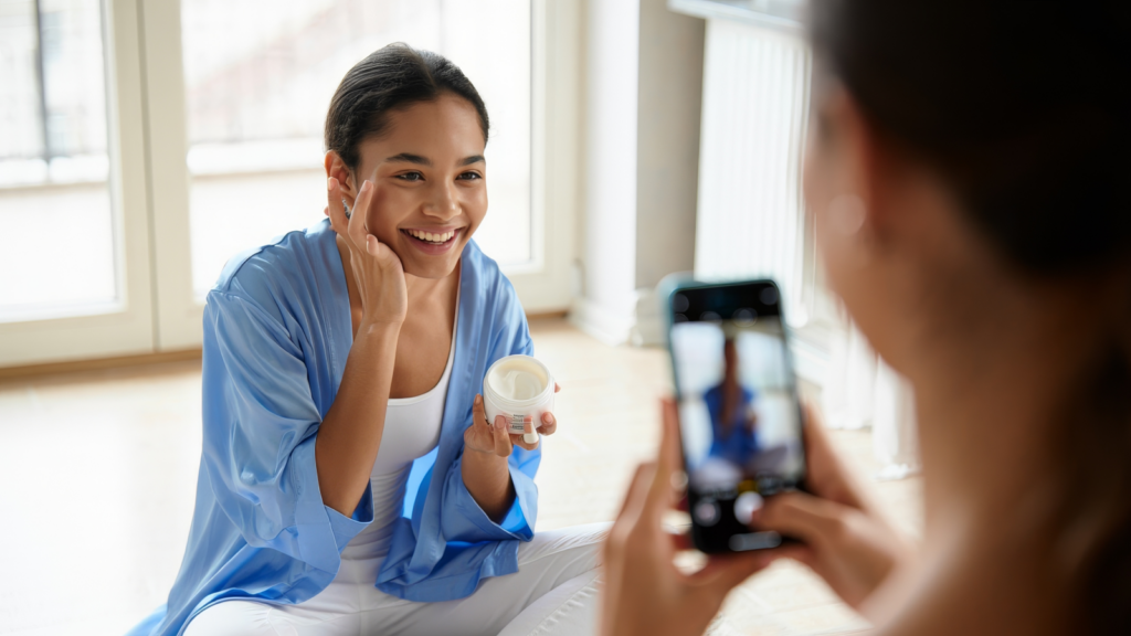 A woman in a blue robe is applying cream to her face, smiling at the camera. Another person photographs her with a smartphone.