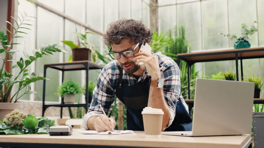 Common mistakes to avoid when getting a business license A man in an apron works on his laptop while speaking on the phone, balancing tasks in a busy environment.