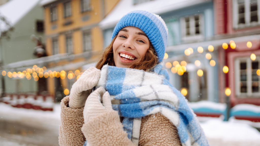 Cozy apparel and warm clothing Smiling woman in winter clothes stands in a snowy street, wearing a blue hat and checkered scarf. Festive lights and houses in the background.