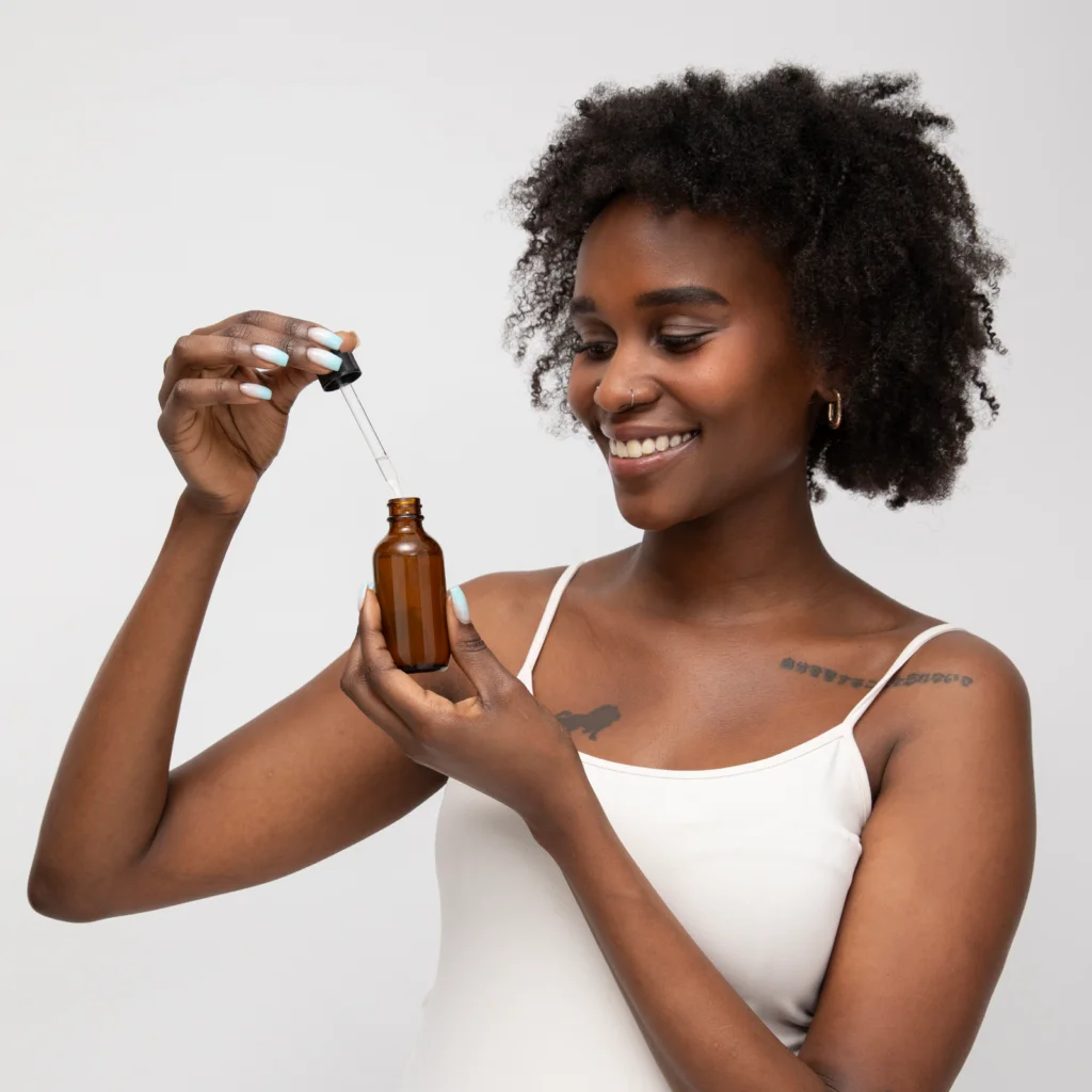 A smiling woman in a white tank top holds a brown bottle with a dropper, suggesting skincare or aromatherapy.