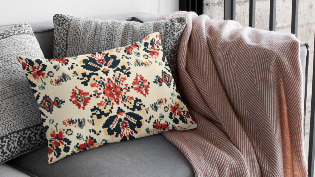 Cross cultural and global inspired prints A patterned pillow with red, navy, and cream designs on a grey couch, alongside a textured grey pillow and a draped blanket.