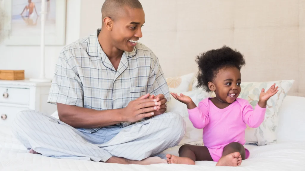 A father and toddler sit on a bed in light pajamas. The father claps while the child raises their hands, both smiling.