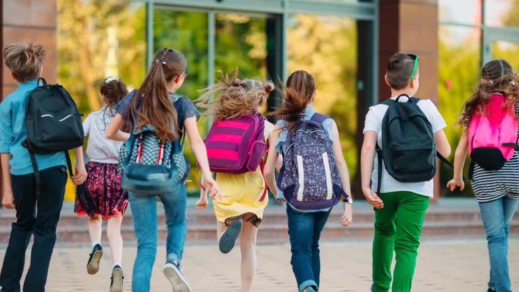 A group of children with colorful backpacks joyfully runs towards the school doors on a sunny day.