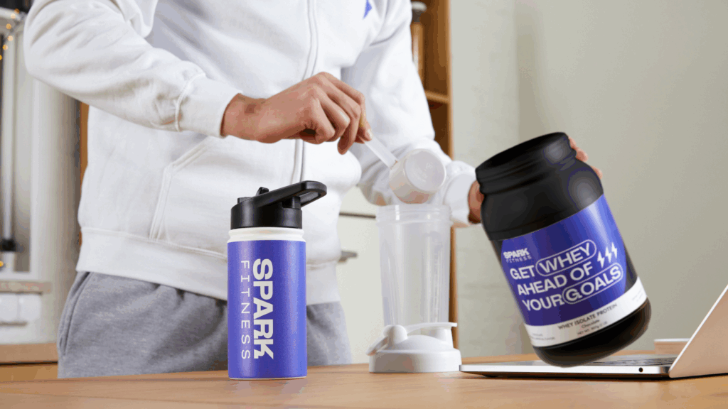 A man in a white hoodie prepares a protein shake, holding whey protein and a scoop over a bottle labeled "Spark Fitness".