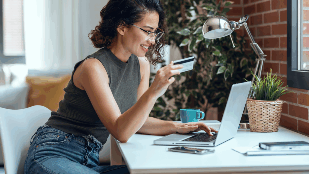 A woman smiles at a laptop, holding a credit card, as she sets up an online store to sell coffee using print-on-demand.