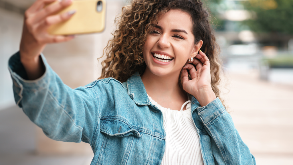 A woman with curly hair smiles while taking a selfie with a smartphone. She wears a denim jacket and stands outdoors.