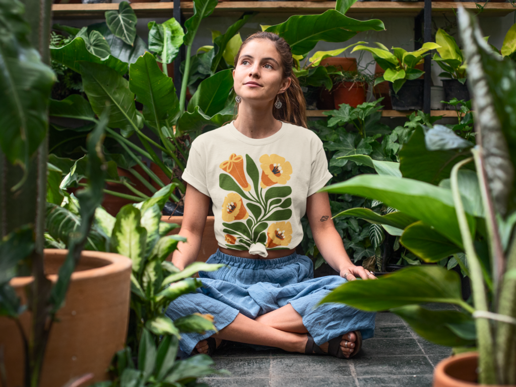 Eco biophilic and nature inspired visuals A woman sits cross-legged in a lush greenhouse, surrounded by vibrant plants. She wears a floral t-shirt and blue skirt.