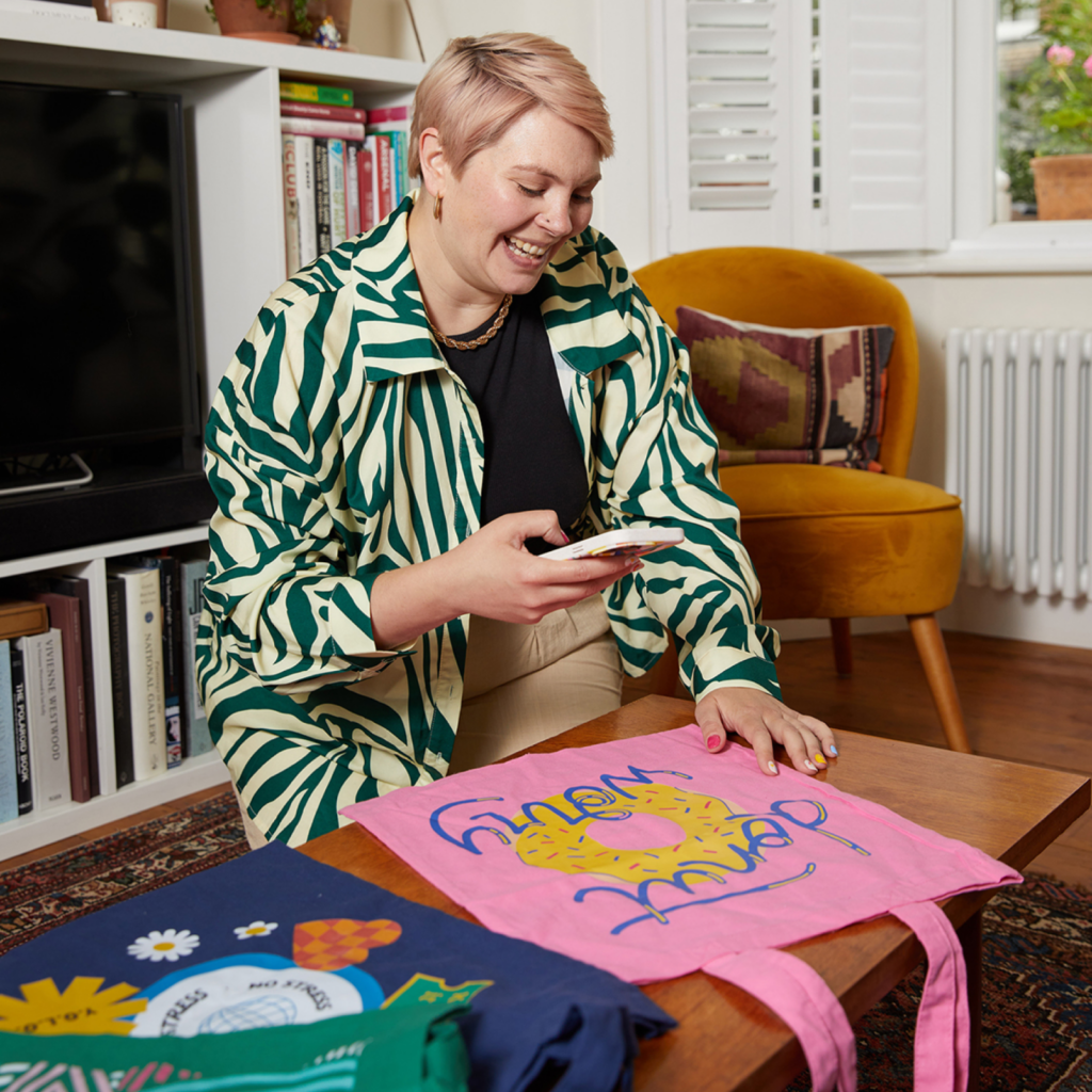 A woman with short hair, wearing a green-striped shirt, smiles while taking a picture with her phone of colorful tote bags on a wooden table.