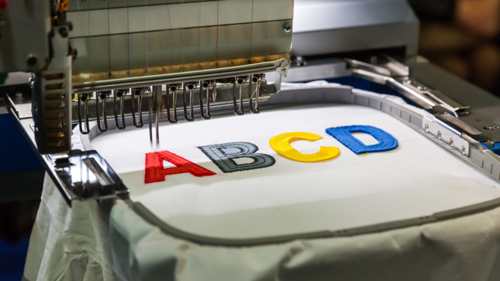 Embroidery machine stitching letters "A B C D" in red, gray, yellow, and blue thread on white fabric.