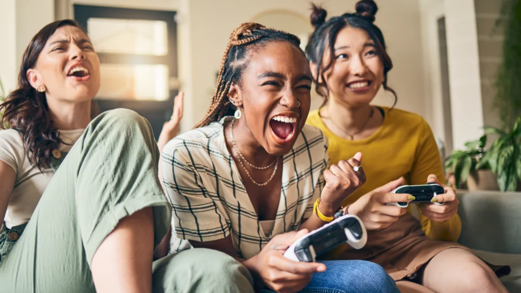 Three women joyfully laughing together while playing video games in a cozy living room setting.