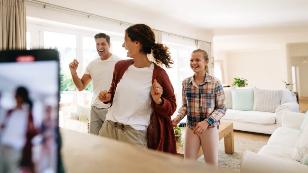A joyful family dances in a bright, modern living room. A smartphone on a small table records their fun.