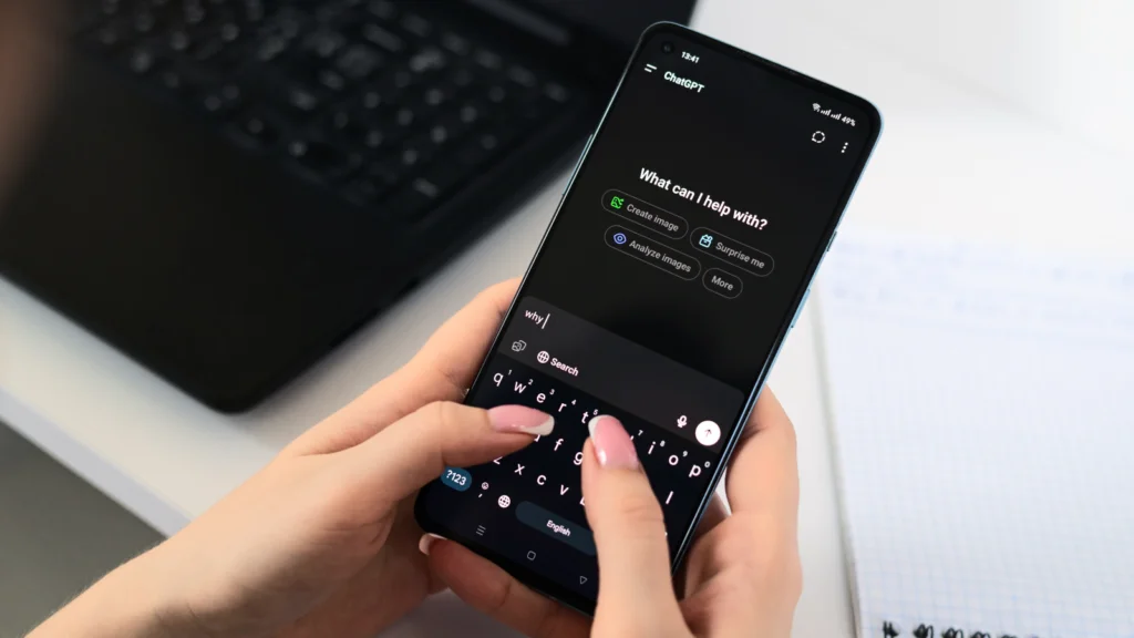 How AI actually predicts viral design trends A person uses a smartphone with ChatGPT open, typing a query on a dark screen. A laptop and a notepad rest nearby.