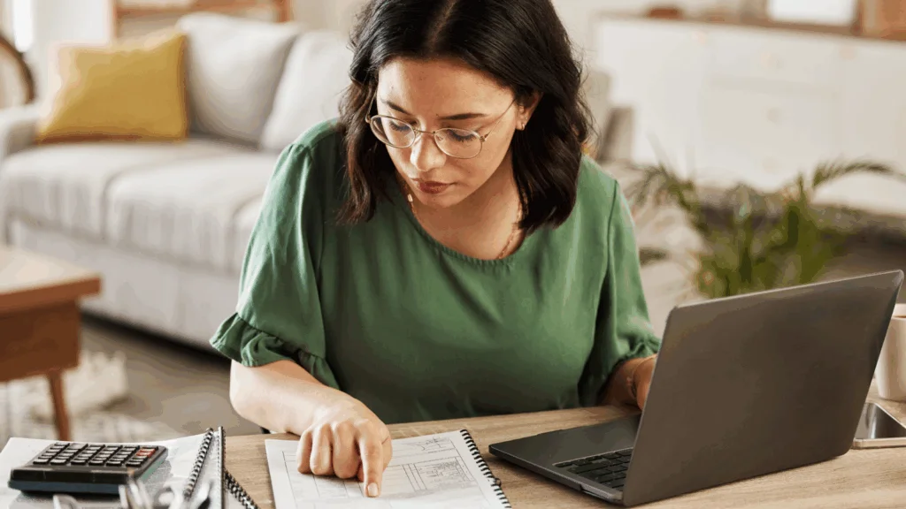 A woman in glasses and a green shirt focuses on paperwork at a home office desk with a laptop and calculator nearby.