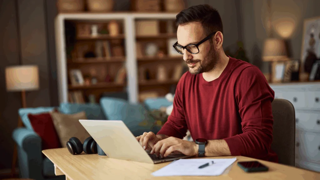 A man wearing glasses and a red sweater types on a laptop at a wooden desk, searching for clothing brand name ideas.