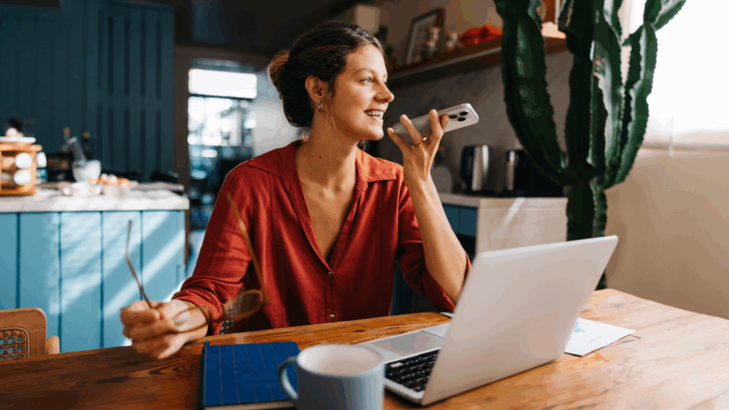 A woman in a red shirt sits at a wooden table, holding a phone and smiling as she uses a laptop to sell coffee online.