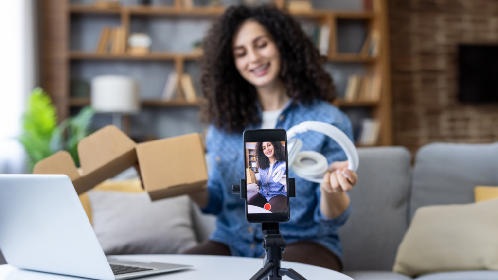 A woman with curly hair is recording herself on a smartphone, unboxing headphones with a joyful expression.