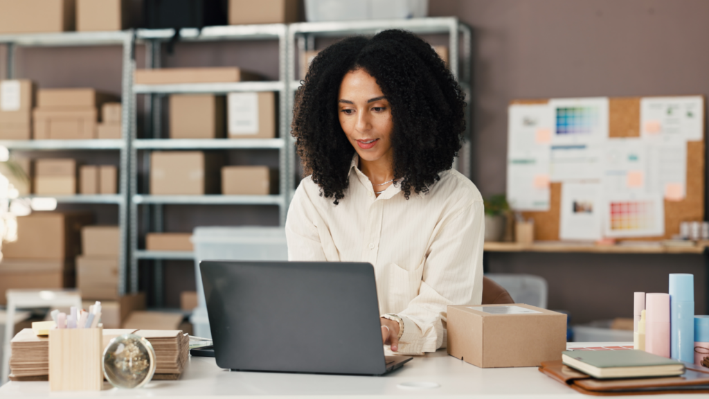 Woman with curly hair working on a laptop in a home office surrounded by shelves of boxes.