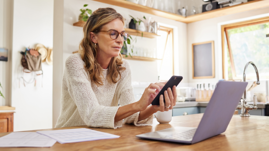 A woman in glasses is sitting at a desk with a laptop, focused on a mobile phone in her hands. Papers scattered around.