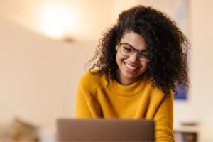 A smiling woman in glasses and a yellow sweater is using a laptop to learn how to use Printify’s Product Creator.