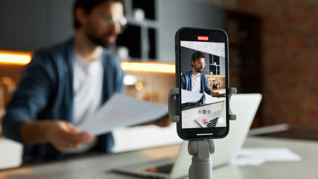 Instagram Reels bonuses and creator programs A smartphone mounted on a tripod records a man in a modern kitchen holding papers with a laptop in front of him.
