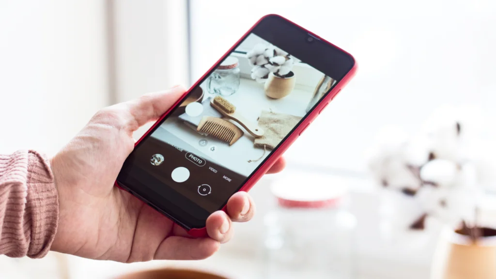 Instagram Reels monetization outlook A hand holds a smartphone with a red case, capturing a photo of a wooden comb, jar, and cotton balls on a windowsill.