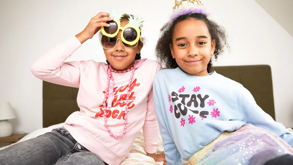 Two smiling kids sitting on a bed. One wears pineapple sunglasses and a pink necklace. The other has a tiara. Both wear colorful shirts.