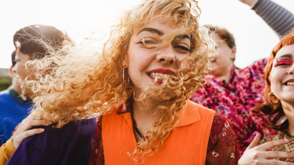Lightweight apparel and festival fashion A joyful group of people with a woman in front wearing bright orange, smiling, with windswept curly hair.