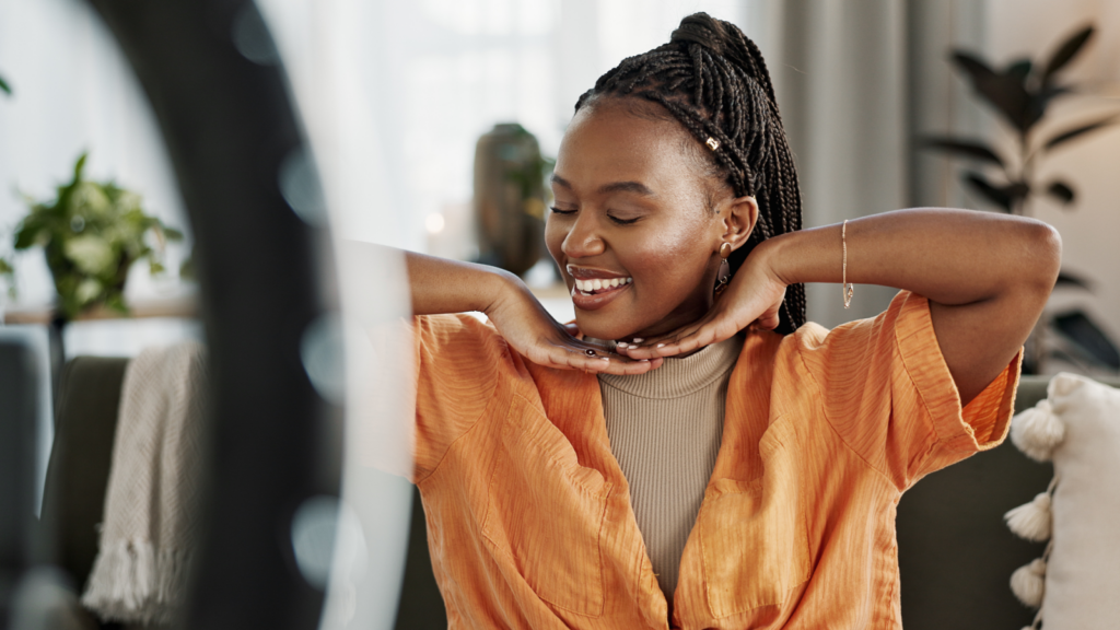A woman with braided hair smiles gently, wearing a bright orange top. She rests her chin on her hands.