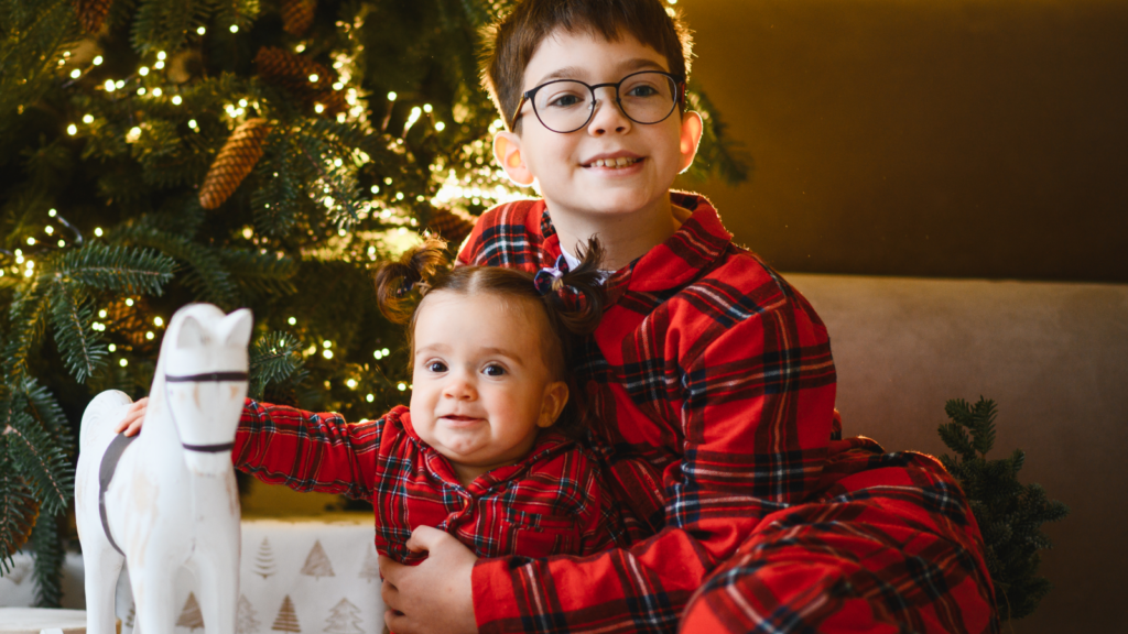 Two children in matching red plaid pajamas sit in front of a Christmas tree. The older one, in glasses, smiles, holding the younger.