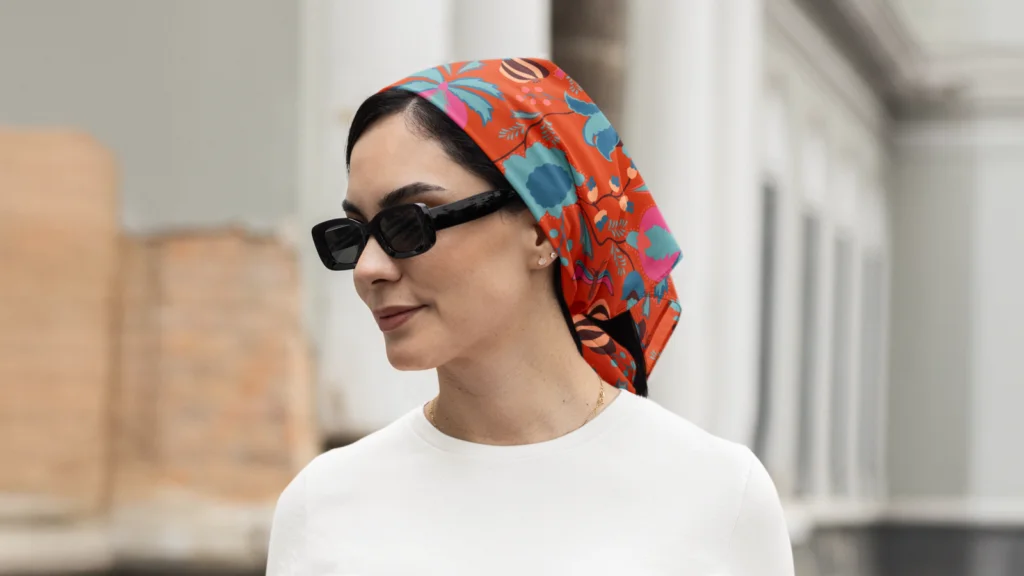Maximalist graphics and loud prints A woman in sunglasses and a vibrant red headscarf with floral patterns stands outdoors against a blurred background, wearing a white top.